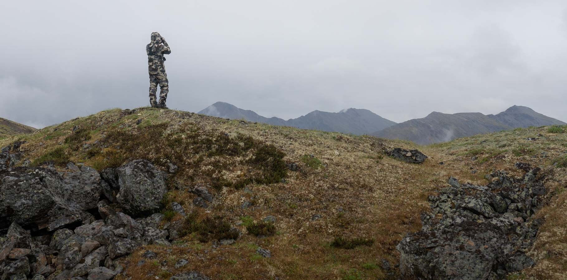 Hunter in Vias Camouflage peering through binoculars over the ridgeline into the distance