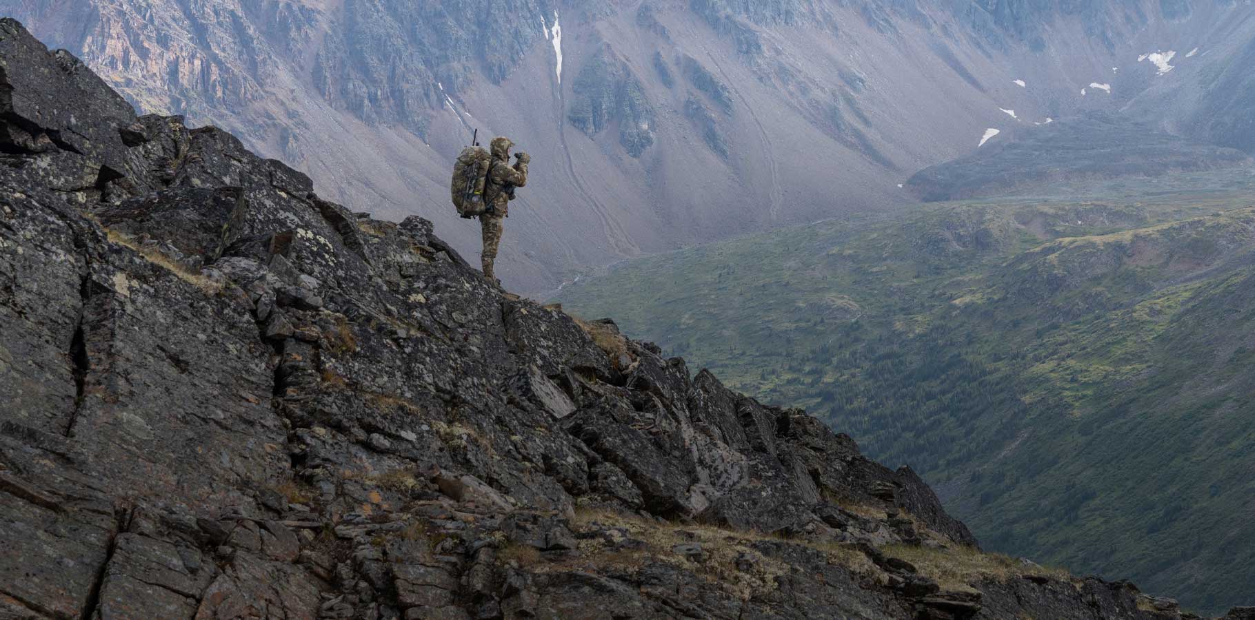 Brendan Burns silhouetted against the distant mountain side while glassing for Stone Sheep