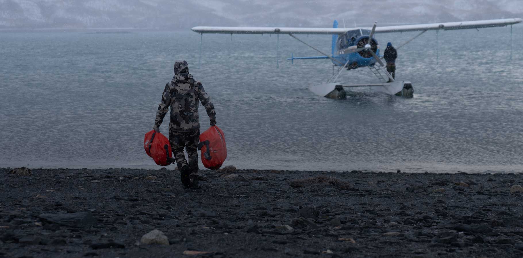 Hunter walking down beachhead towards float plane while carrying two blaze orange taku duffels