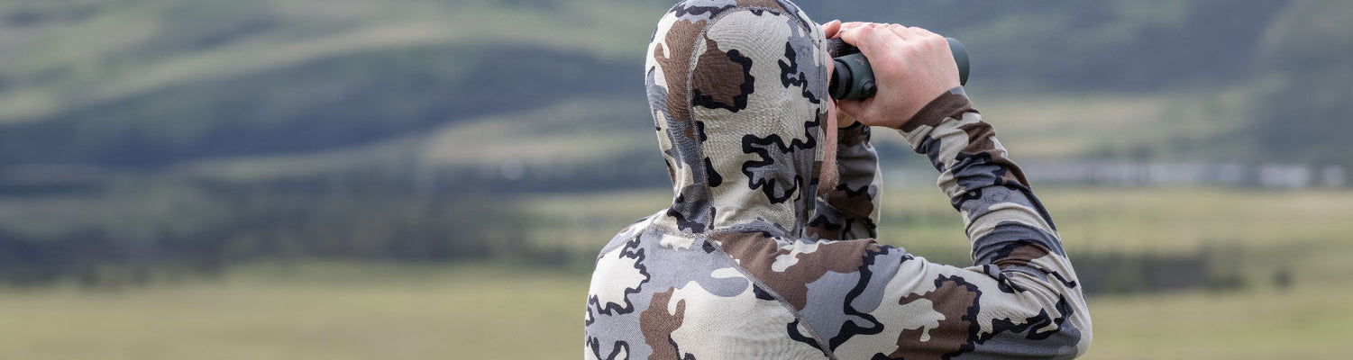 Hunter in an outdoor setting, using a binocular, wearing a PRO Merino 200 Zip-T Hoodie in Vias Camouflage