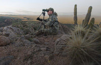 Hunter in Valo camouflage seated and looking through binoculars mounted to tripod