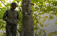 Bowhunter in ash brown Encounter vest and Verde camouflage Encounter pant standing in a treestand with bow hanging beside him