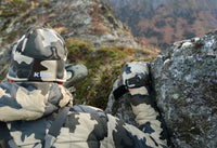 Hunter in Vias camouflage peering over edge of rocks while looking through spotting scope