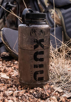 Well worn KUIU 32oz Wide Mouth Bottle on ground with partial shot of a pack in the background