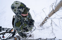 Bird's view of Hunter standing in a tree stand above the ground covered in snow, with his bow and arrow by his side