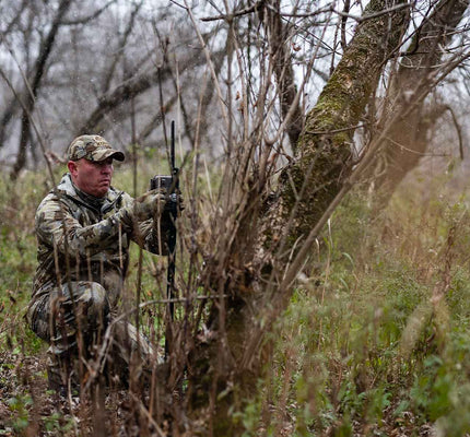 Hunter in Valo camouflage squatting behind a tree in wet conditions